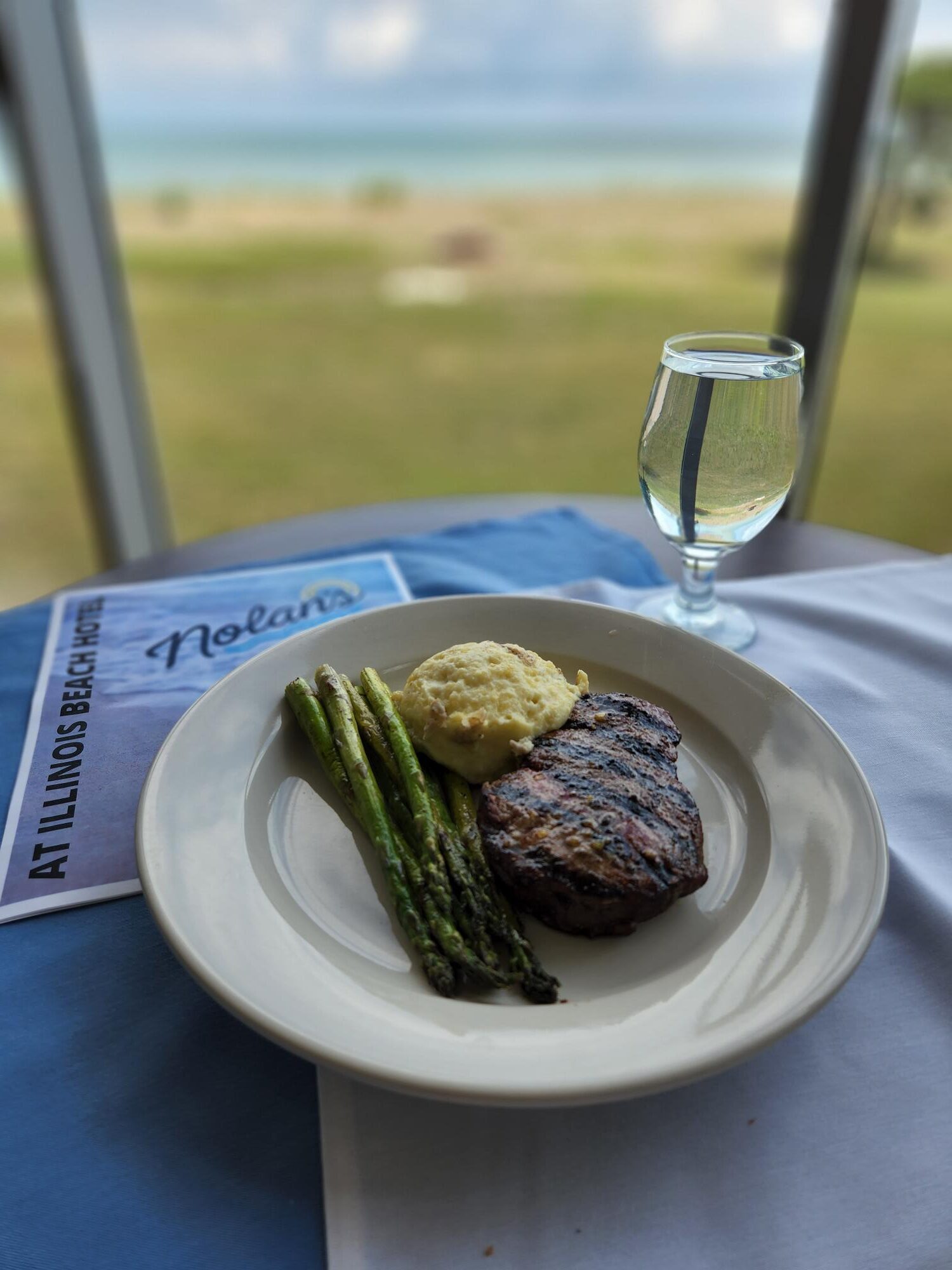 Steak, potatoes and asparagus on white plate with a glass of water.